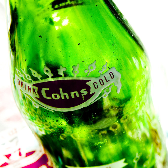 Green glass bottle, slightly tipped on the side to showcase the label that sits across the top of the ridge, the label reading 'Drink Cohn Cold'.