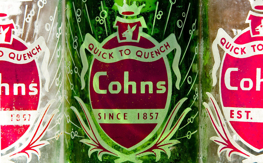 Three glass bottles in a row, two clear and the one in the middle green glass. They all have labels in red and white colouring and the saying 'quick to quench'.