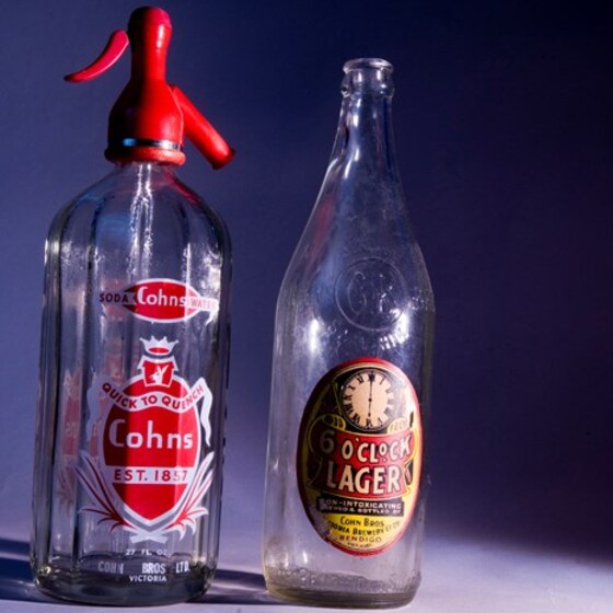 One glass bottle and one larger glass syphon with a red nozzle on top. Both have labels on the front in different colours and with different detailing.