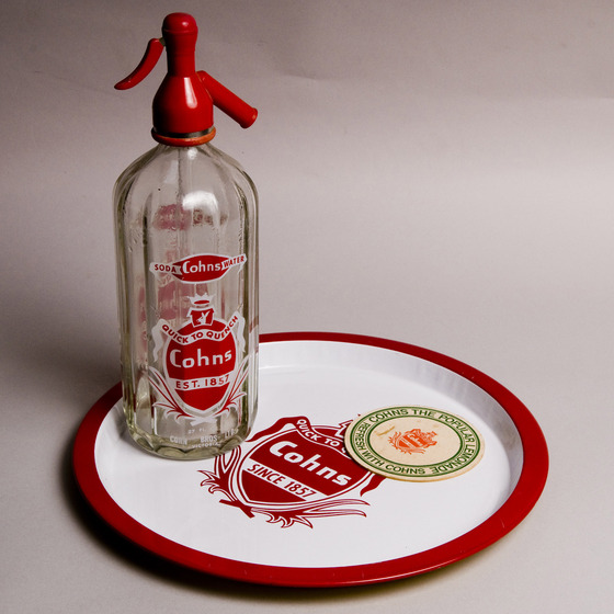 A glass bottle with a red nozzle on top sits on a white and red round tray. Both the bottle and the tray have a ed and white Cohns label on them. The tray also has a round paper coaster sitting on top.