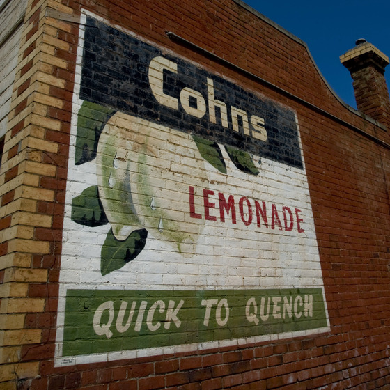 A large sign on the side of a brick building. The sign is broken into thirds, the top a blue band with the word 'Cohns', the centre third with a picture of hanging lemons and the word ' lemonade', and the bottom third a green banner with the text 'Quick to Quench'.