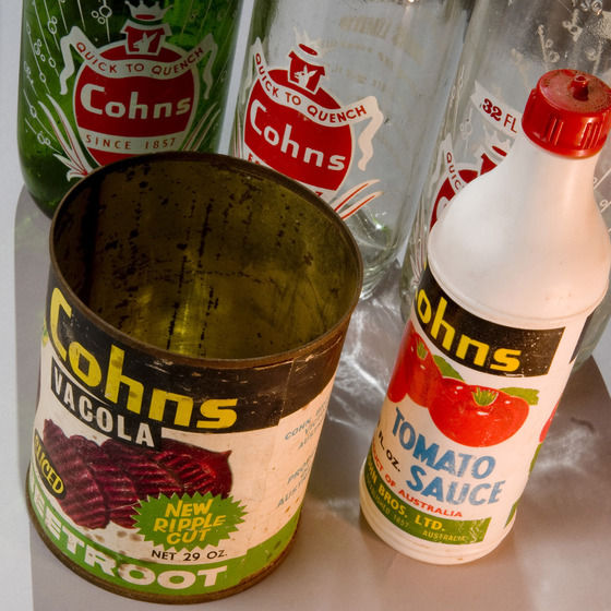 A large round empty tin, a tall white bottle, and three glass bottles, all with different labels printed on the front promoting the Cohns company. The tin includes a picture of beetroot slices and the white bottle a picture of tomatoes.