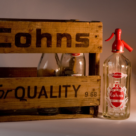 Wooden crate with slats going both horizontal and vertical. Printed on the side in black text is 'Cohns for Quality'. In front of the box is a glass syphon with a red nozzle, and within the crate are two glass bottles.