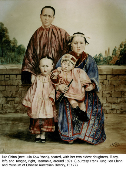 A Chinese family sit and stand in front of a short brick wall. One woman stands at the back in a long brown robe, her hand on the shoulder of a woman sitting in a decorative blue and red robe. The sitting woman has a small child sitting on her knee in a knee length pink robe and decorative hat, whilst another child leans on her in a pink robe, red pants and her hair pulled back tight off her face.