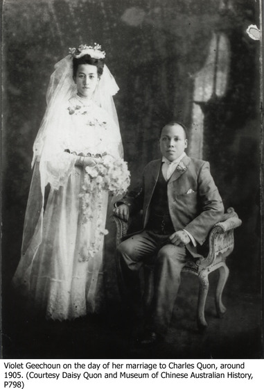 A formal portrait of a bride and groom. The bride stands in a floor length, and wrist length, white gown with a head piece and veil trailing behind her. She is holding a small bouquet of flowers. The groom sits in a decorative chair beside her, dressed in a three-piece suit.