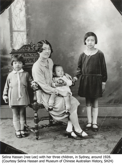 A family pose for a portrait in a room of a house. The mother sits on an ornate wooden chair, a toddler sitting on her knee. Two other children stand either side of the chair, both in knee length dresses, pulled up socks and black shoes.