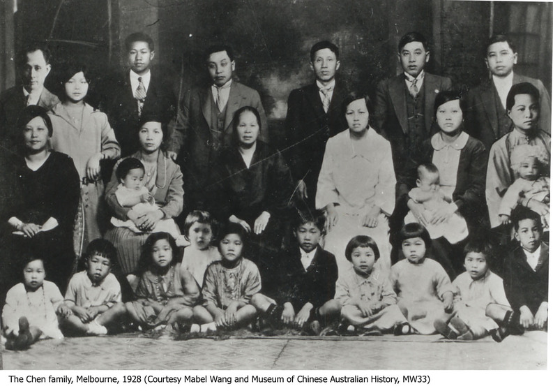 A large family portrait, made up on three rows of people. The back row, made up of men except for one woman, stand in front of a row of seated women. At the feet of the women are children sitting cross legged on the floor.