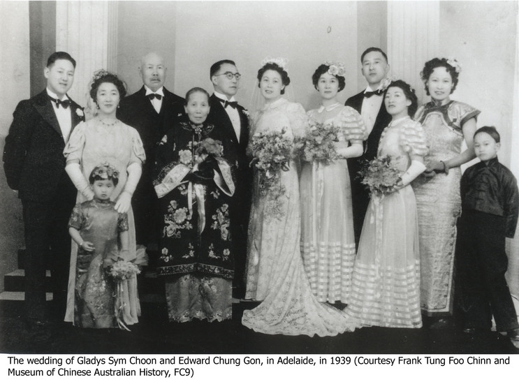 A formal wedding party group portrait. The bride and groom are positioned in the centre of the group, with the guests around them dressed in formal suit and floor length gowns, as well traditional Chinese outfits on an older lady and two small children.