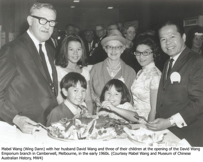 A group photo at the opening of a new store. Five people stand in the back row, both men and women, whilst two children stand in front of them, picking at a plate of food on a table. The entire group is smiling and looking at the camera.