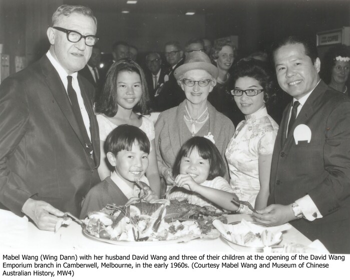 A group photo at the opening of a new store. Five people stand in the back row, both men and women, whilst two children stand in front of them, picking at a plate of food on a table. The entire group is smiling and looking at the camera.