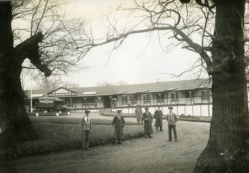 Black and white photograph of a group of men in uniform standing in front of a long pavilion. Two large trees frame the men and in the background a hospital truck can be seen parked outside the pavilion.