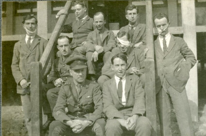A group of men in suits sit and stand next to a set of wooden verandah stairs. Some of the men have bandages on their face in various positions, while others visible physical war damage can be seen around their heads.