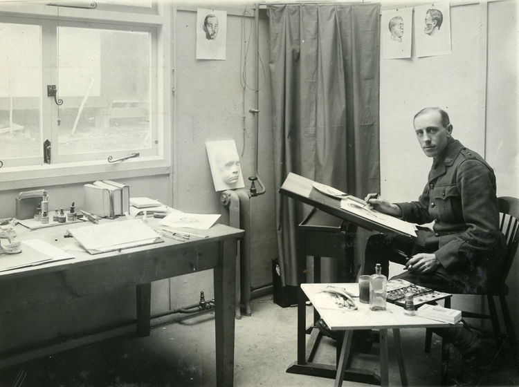 A man sits at a desk, the top sloping towards him with a hand poised over a piece of paper, pen in hand. At a small table next to him is a paint palette and bottles of liquid. He is staring at the camera but around him are drawings of deformed faces and a plaster cast sitting directly in front of his table.