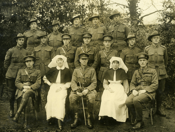 Posed photograph of a group of men dressed in Australian military uniforms and two women in nurse uniforms. They're lined up in three rows in a garden setting.