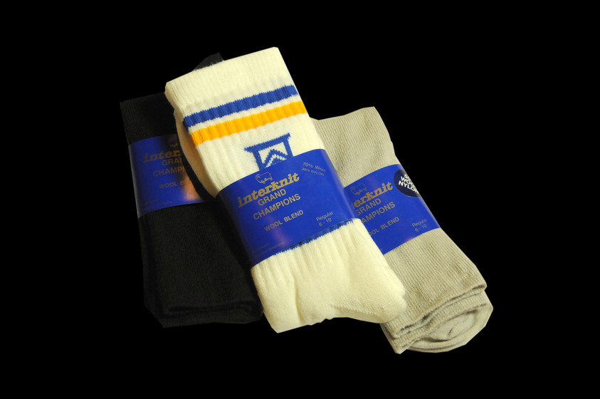Three pairs of socks stacked on top of one another. The top pair are yellow in colour with a blue and yellow band around the ankle. The bottom left pair is black and the bottom right pair an olive green. All pairs are wrapped in a blue cardboard band.