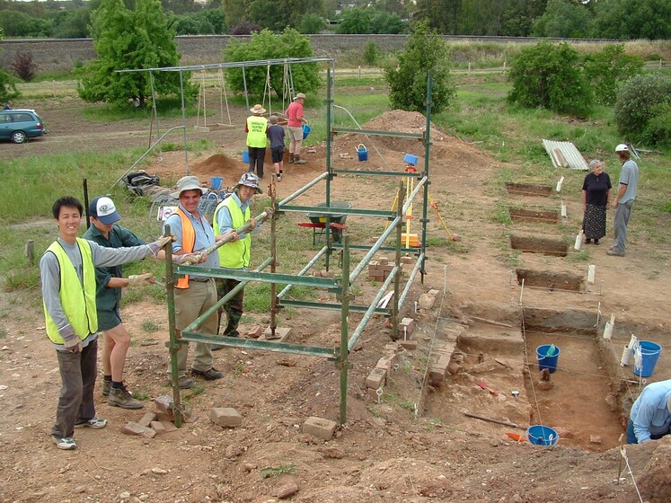 A group of people in high vis stand in a dirt patch, holding up a metal structure that is positioned over a rectangular area of soil. Behind to the left are more people walking away from the dig site, and to the right is an older woman and man standing next to a set of carved out steps in the earth.