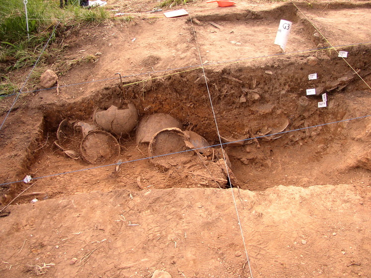 Shards of clay pots sit partially submerged in a dirt dug out. String is positioned in squares over the top of the dug out and small white tags are positioned in various places around the site.