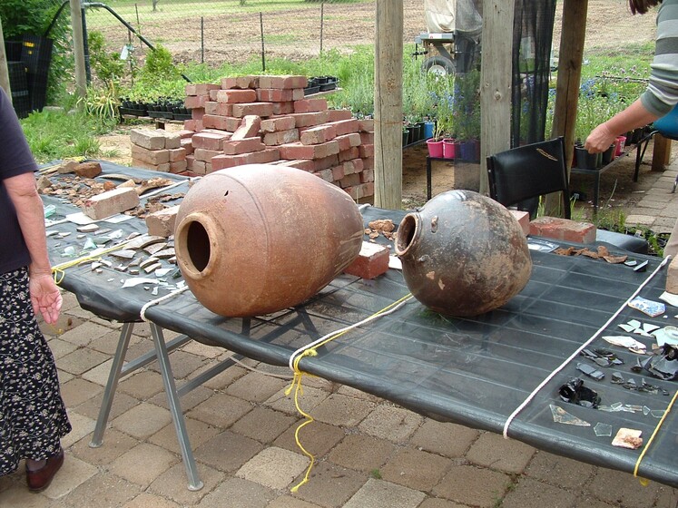 Two large cylindrical pots sit on their sides on a table made of a black mesh. Around the pots are shards of different colours and sizes, grouped into sections.