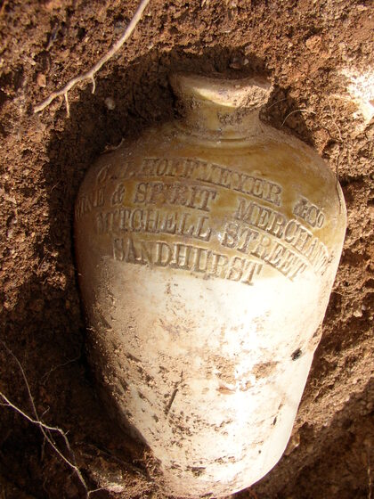 A large demijohn lays partially submerged in soil. Around the neck of the object is inscribed text, reading 'O.H. Hoffmeyer & Co. Wine & Spirit Merchant Mitchell Street Sandhurst'.