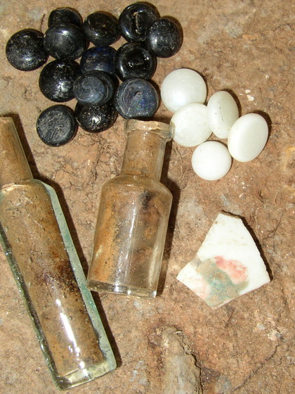 Different groups of artefacts lie on a dirt patch. They include broken glass bottles, ceramic shards and glass stones.