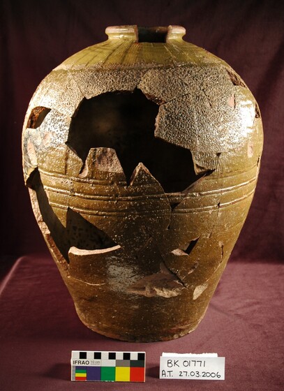 A broken ceramic jar stands upright on a burgundy table cloth covered table. There are large parts of the jar missing, but certain parts have been joined back together. A colour matching strip and catalogue number are places in front of the jar.
