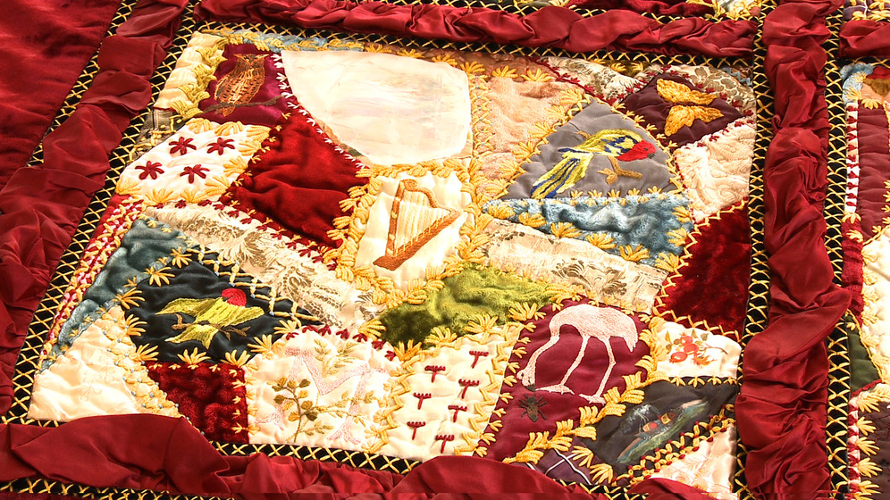 A panel on a patchwork quilt, made up of various pieces of different coloured fabric and embroidered images and stitches over the top.