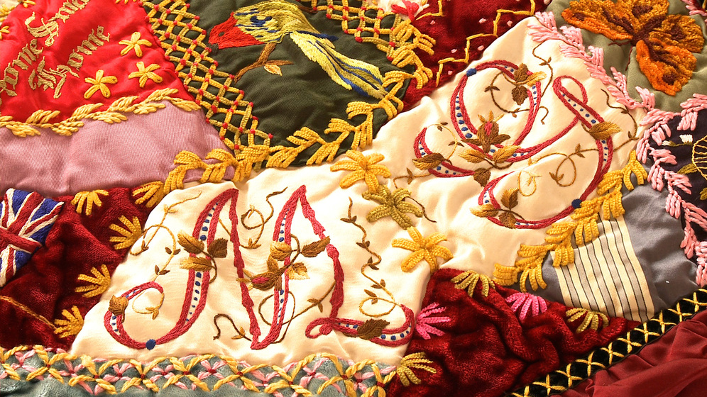 A panel on a patchwork quilt, made up of various pieces of different coloured fabric and embroidered images and stitches over the top, including the initials M and G.