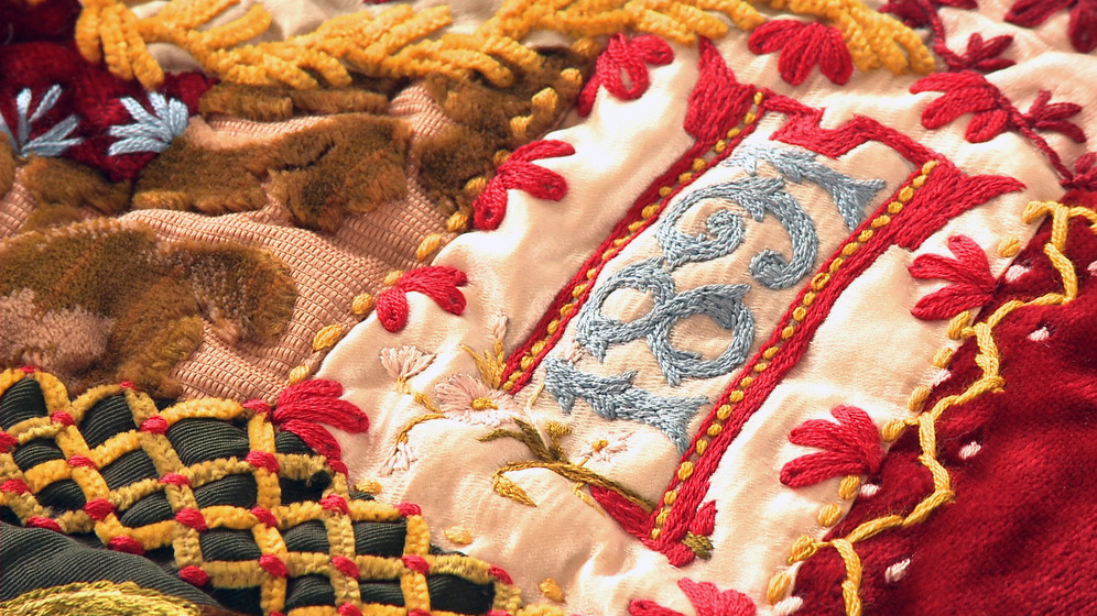 A panel on a patchwork quilt, made up of various pieces of different coloured fabric and embroidered images and stitches over the top, including the text '1891'.