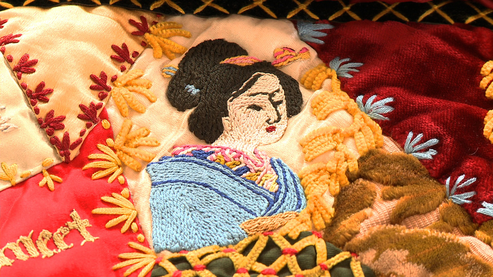 A panel on a patchwork quilt, made up of various pieces of different coloured fabric and embroidered images and stitches over the top, including a detailed image of a geisha woman in a blue robe.