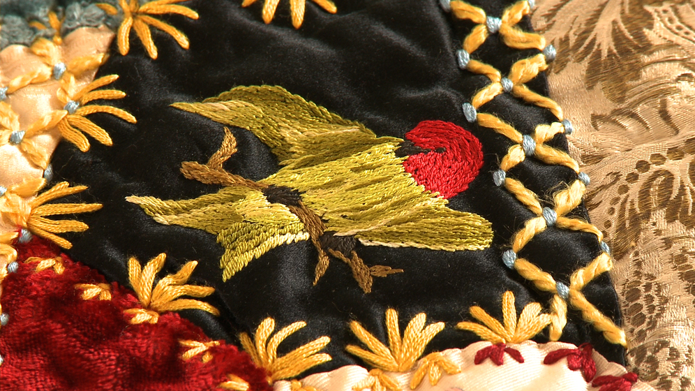 A panel on a patchwork quilt, made up of various pieces of different coloured fabric and embroidered images and stitches over the top, including a detailed image of a red and gold parrot.