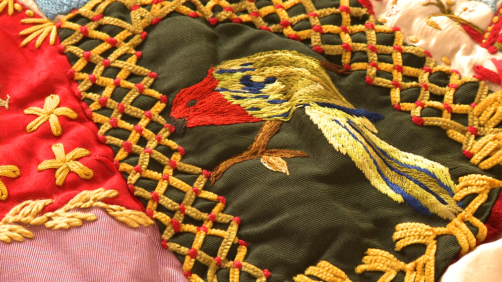 A panel on a patchwork quilt, made up of various pieces of different coloured fabric and embroidered images and stitches over the top, including a detailed image of a red, blue and gold parrot.