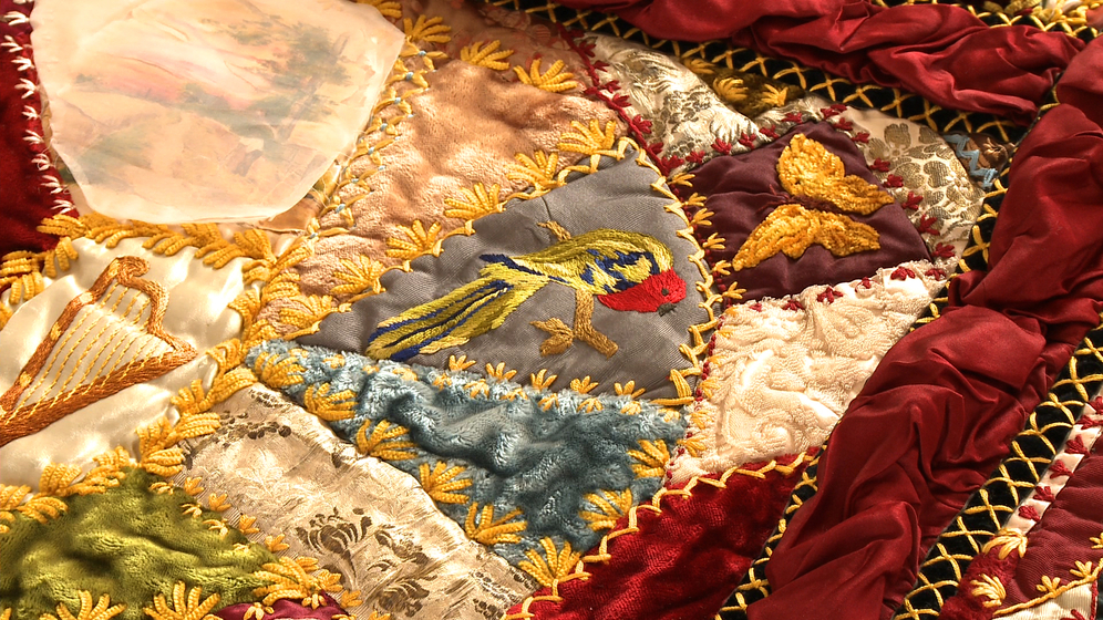 A panel on a patchwork quilt, made up of various pieces of different coloured fabric and embroidered images and stitches over the top, including a detailed image of a red, blue and gold parrot and a golden butterfly.