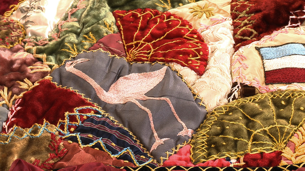 A panel on a patchwork quilt, made up of various pieces of different coloured fabric and embroidered images and stitches over the top, including a pink flamingo with its head tilted up towards the horizon.