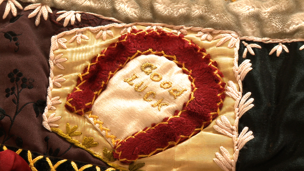 A panel on a patchwork quilt, made up of various pieces of different coloured fabric and embroidered images and stitches over the top, including a detailed picture of a red horseshoe with the words inside 'Good Luck'