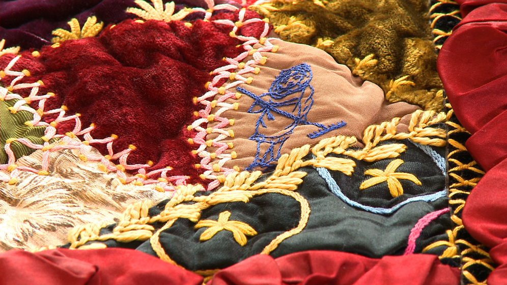 A panel on a patchwork quilt, made up of various pieces of different coloured fabric and embroidered images and stitches over the top, including a detailed image of a girl holding a rake in her hands.