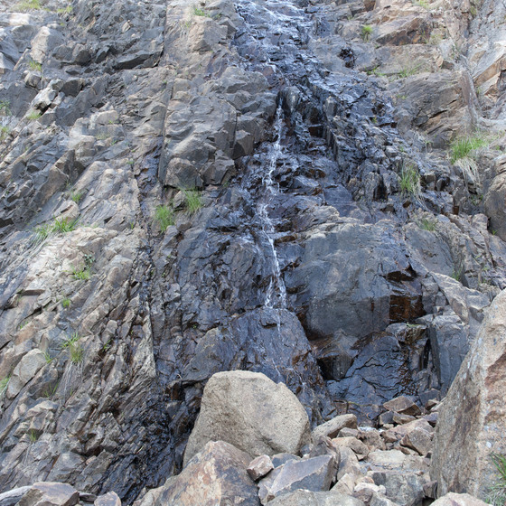 Rocky cliff with moss patches viewed close-up from below. Large dry rocks are in the foreground. The middle of the cliff's rocks are damp, and a light waterfall is running down.