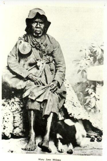 An early black and white photograph of a seated elderly woman wearing a hat, large coat and skirt, with a black and white dog at her feet.