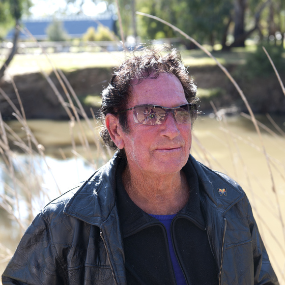 Man in close up wearing leather jacket with a butterfly pin on the left collar, zip up top beneath, and sunglasses. He is standing among tall dried grass with a sunlit field and trees behind. 
