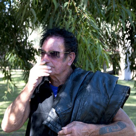 Man in close up wearing a sleeveless zip up vest, sunglasses, and has a leather jacket hung over his left shoulder. He holds his right arm up to his mouth. He is cast in shadow, with gum leaves behind.