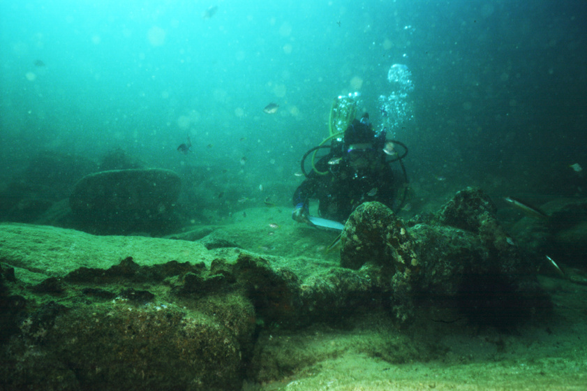 A diver swims very close to a metal artefact sitting on the ocean floor. The artefact is covered in plant life and barnacles.