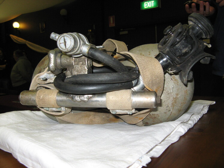 A set of diving equipment sits on a white cloth on a table top. The breathing apparatus and monitor can be seen resting on the tank.