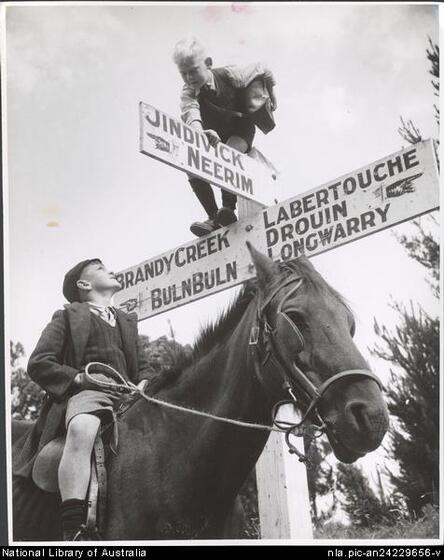 Black and white image of a young boy on a horse wearing a cap, shorts and jacket, looking up at a young boy standing on top of a three way white directional post. Directions are to Brandy Creek and BulnBuln, Labertouche, Drouin and Landwarry, and Jindiwick Neerim.