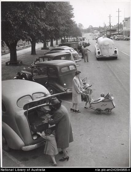 1940s era cars parked diagonal along a busy tree-lined street. A woman in a long cloak and heels and a child in a coat are taking a case out of the front car's book or trunk. Behind a woman in a light long coat and heels pushes a baby in a carriage with a child in a hat at her side.