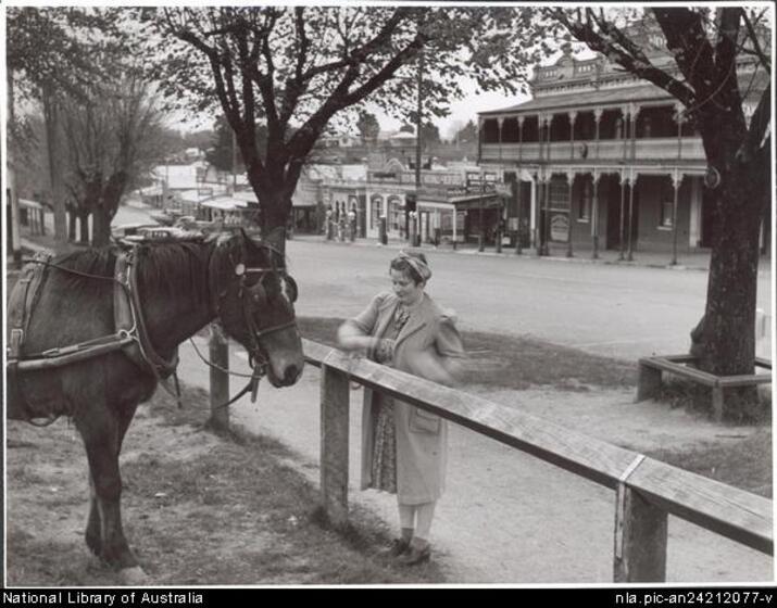 A woman in a long coat, flowered dress and hat ties a horse to a fence post. Trees and a township main street are in the background.