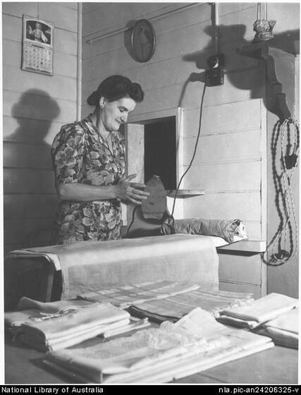 Woman in a floral dress holding her right hand up to the bottom of an iron, and standing behind an ironing board with cloth on it. Fabric is folded in the foreground on a table, and she is in a small weatherboard room with a calendar and plate hanging on the walls.