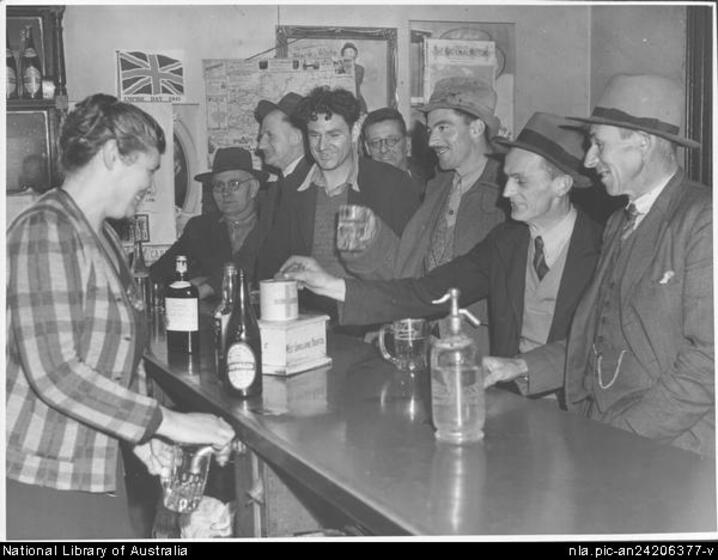 A woman in a checked blazer pours a beer from a tap behind a bar. A group of seven smiling men sit and stand on the other side of the bar. Wine bottles and a seltzer bottle are on the bar top.