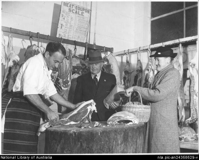 A man in a striped butcher's apron holds a portion of meat on a large tree stump used as a table. An older man and woman in coats and hats with a basket look on, hanging meat is behind with a sign above featuring the heading 'Meat Coupon Scale'.