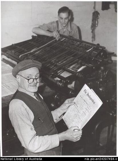 Elderly man wearing glasses, a cap, long sleeved shirt and vest holding a newspaper and standing in front of an printing press operated from behind by a younger man in a shirt and braces.