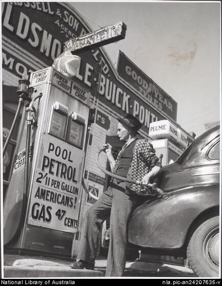 Man in a check cardigan, vest and trousers leans back on the boot or trunk of a car, holding a petrol hose attached to the fuel filller hole of the car. He is looking away from the car at the petrol pump featuring the words 'pool petrol 2'11 per gallon Americans! gas 47 cents'.