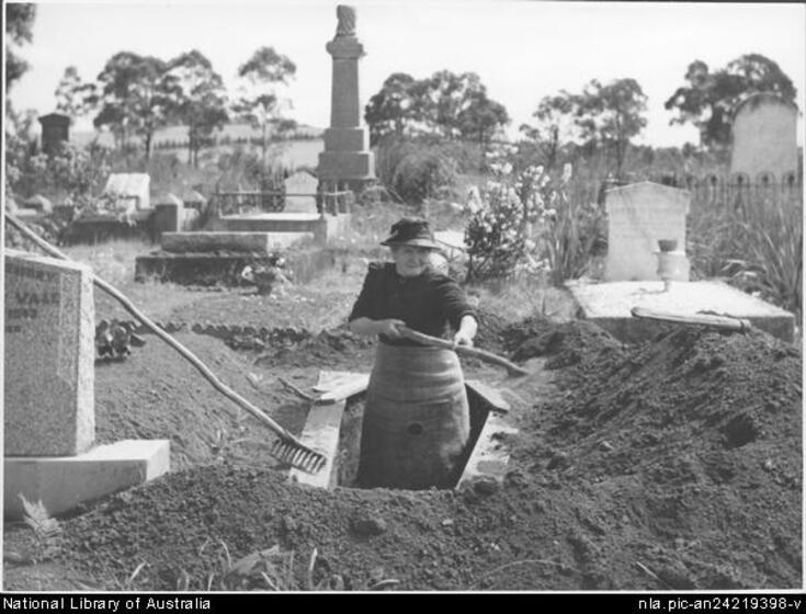 Middle aged woman in a shirt, black long sleeved top and wide brim hat stands in an overgrown cemetery in a dug grave holding a stick. A pile of dirt is to her left. 
